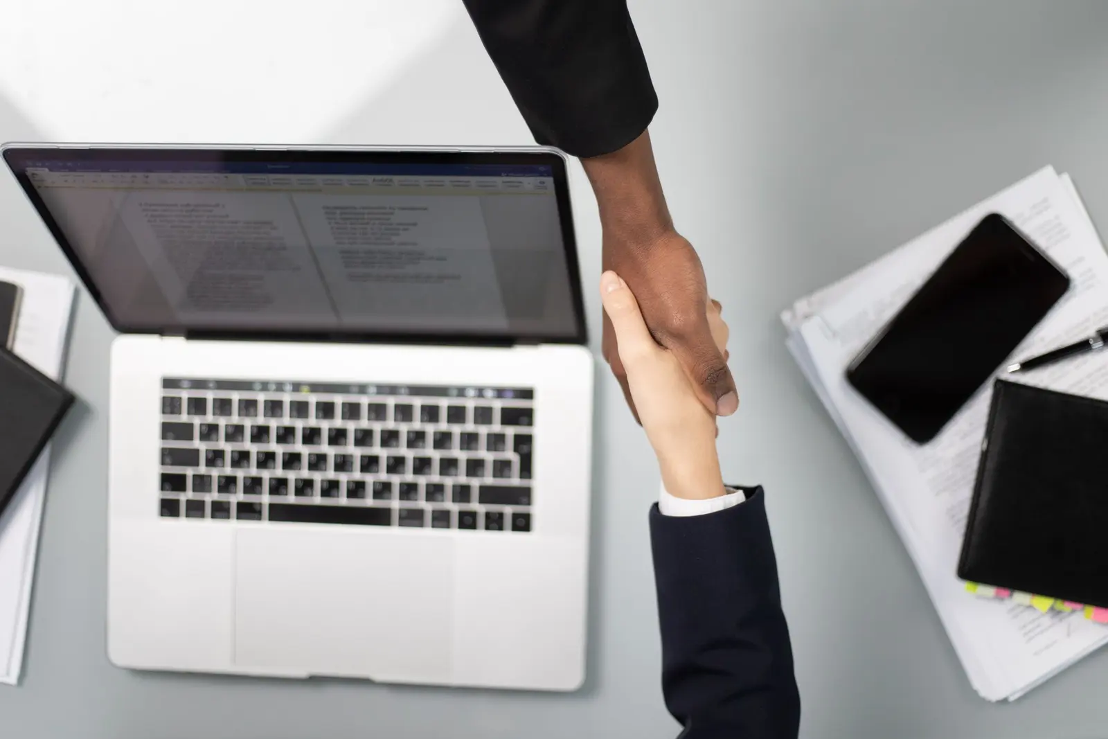 Two people shaking hands above a laptop and project documents